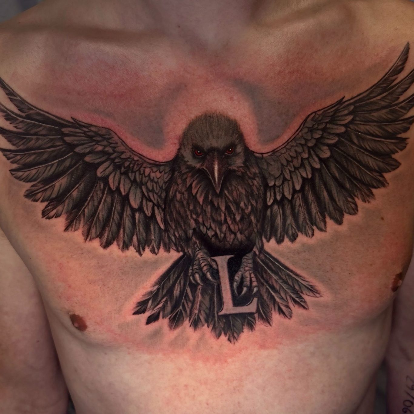 Tätowierung eines großen, fliegenden Raben mit dem Buchstaben "L" auf der Brust.