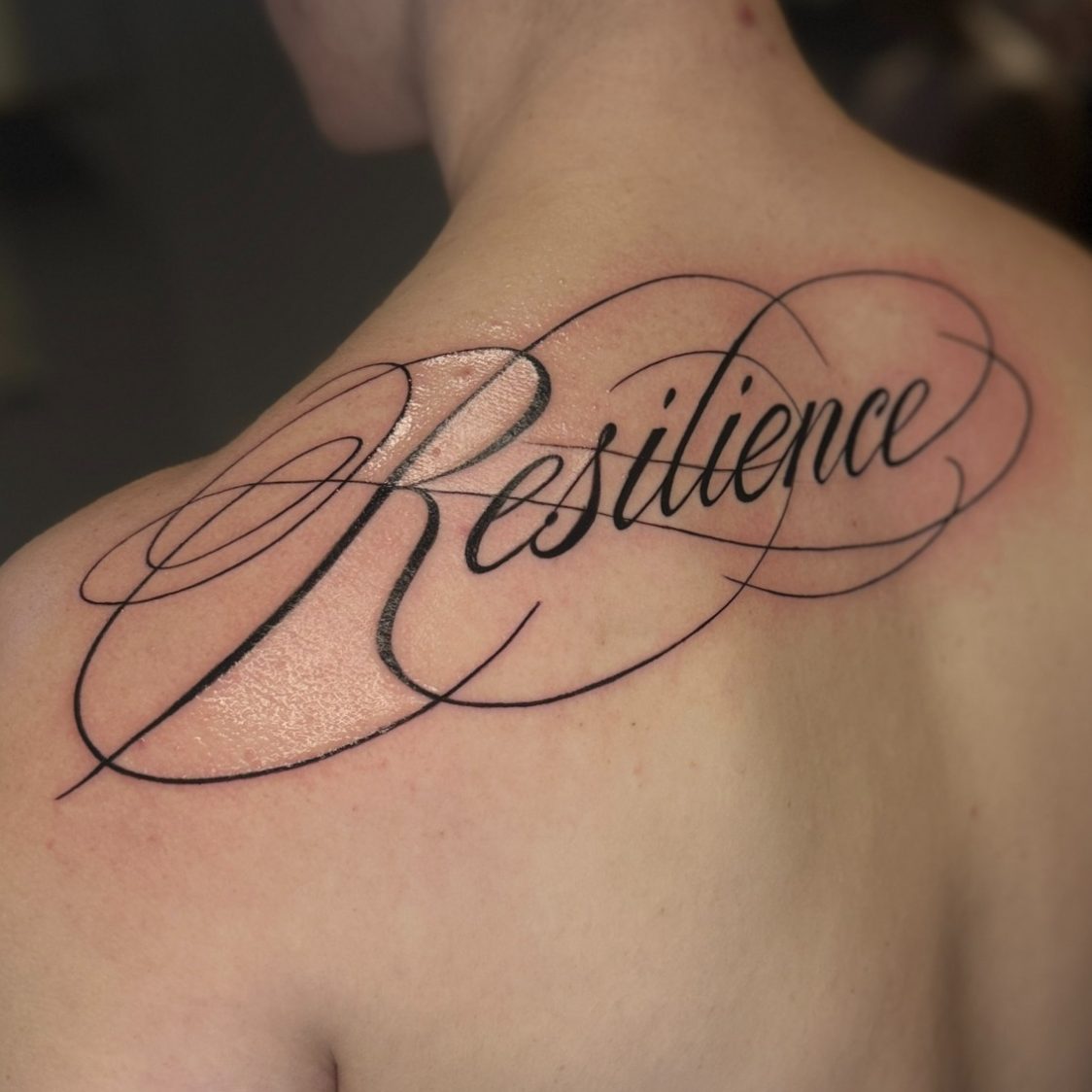 Fineline Lettering Tattoo auf dem Rücken mit dem Wort "Resilience" in geschwungener Schrift.